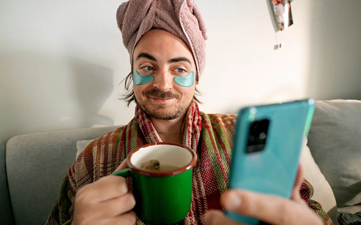 Ein Mann sitzt mit Augenpads und Handtuch auf dem Kopf auf der Couch und schaut zufrieden auf sein Smartphone