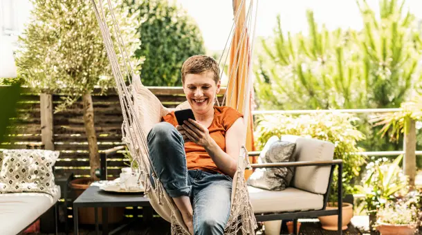Frau sitzt lächelnd in einer Hängematte und guckt auf ihr Smartphone.