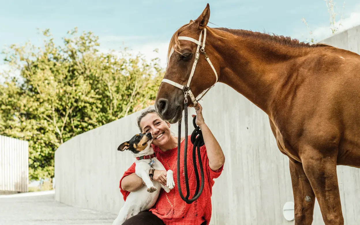 Eine Frau kniet lachend auf dem Boden und hält einen Hund sowie ein Pferd.