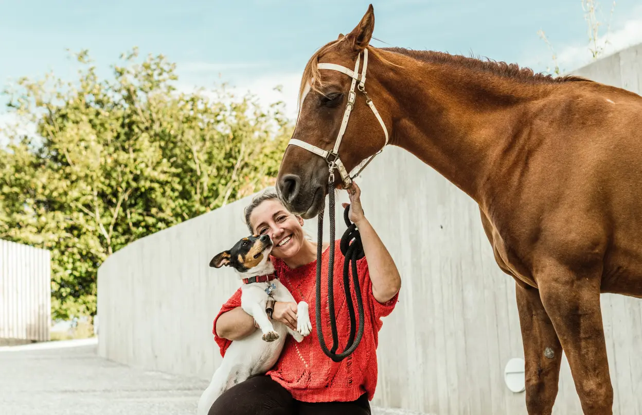 Eine Frau kniet lachend auf dem Boden und hält einen Hund sowie ein Pferd.