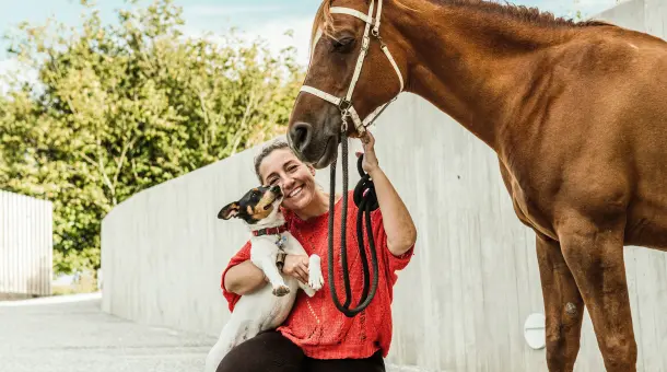 Eine Frau kniet lachend auf dem Boden und hält einen Hund sowie ein Pferd.