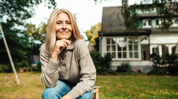 Eine Frau sitzt fröhlich im Garten vor einem Haus.