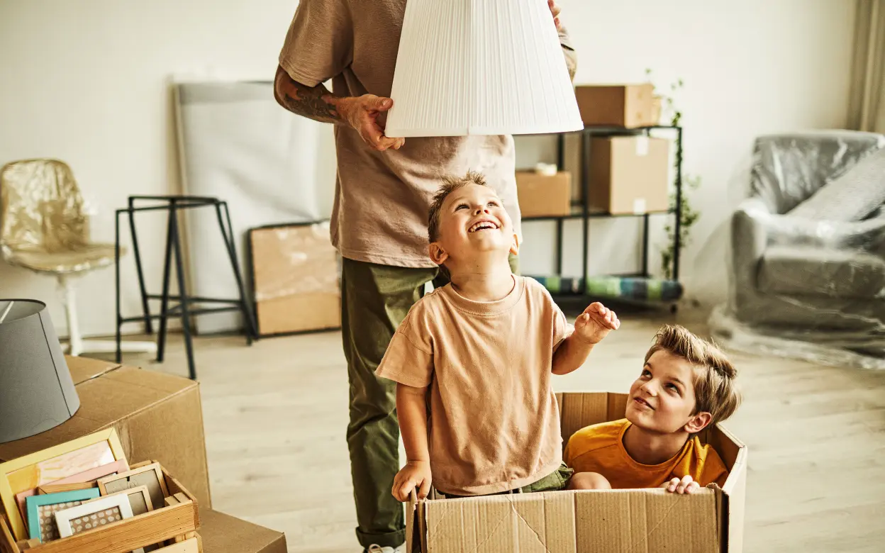 Mann hält Lampenschirm über Kind in Umzugskarton.
