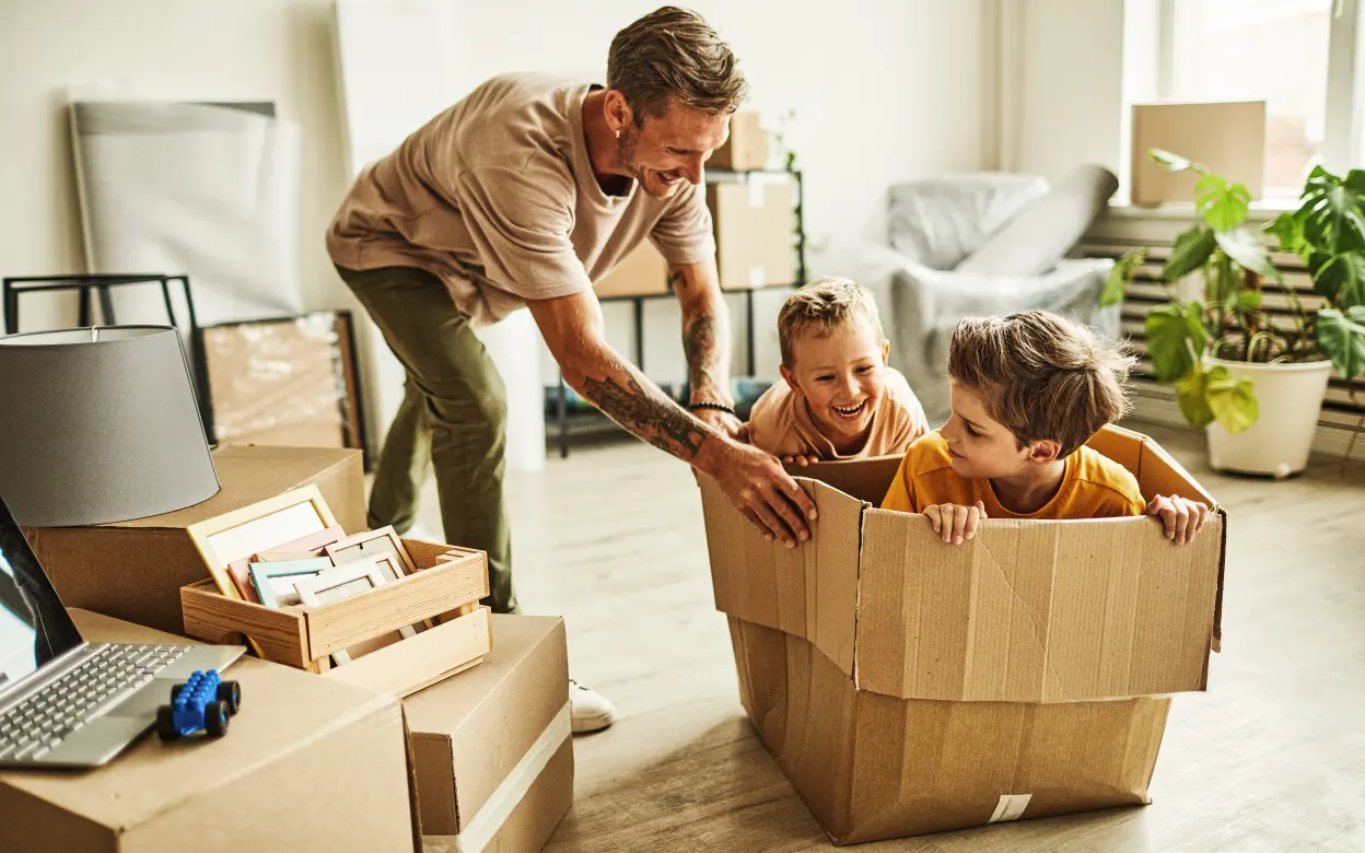 Ein Mann schiebt zwei Kinder in einem Umzugskarton durch ein Zimmer.