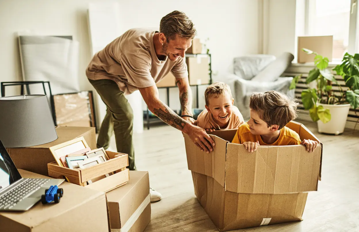 Ein Mann schiebt zwei Kinder in einem Umzugskarton durch ein Zimmer.