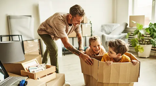 Ein Mann schiebt zwei Kinder in einem Umzugskarton durch ein Zimmer.