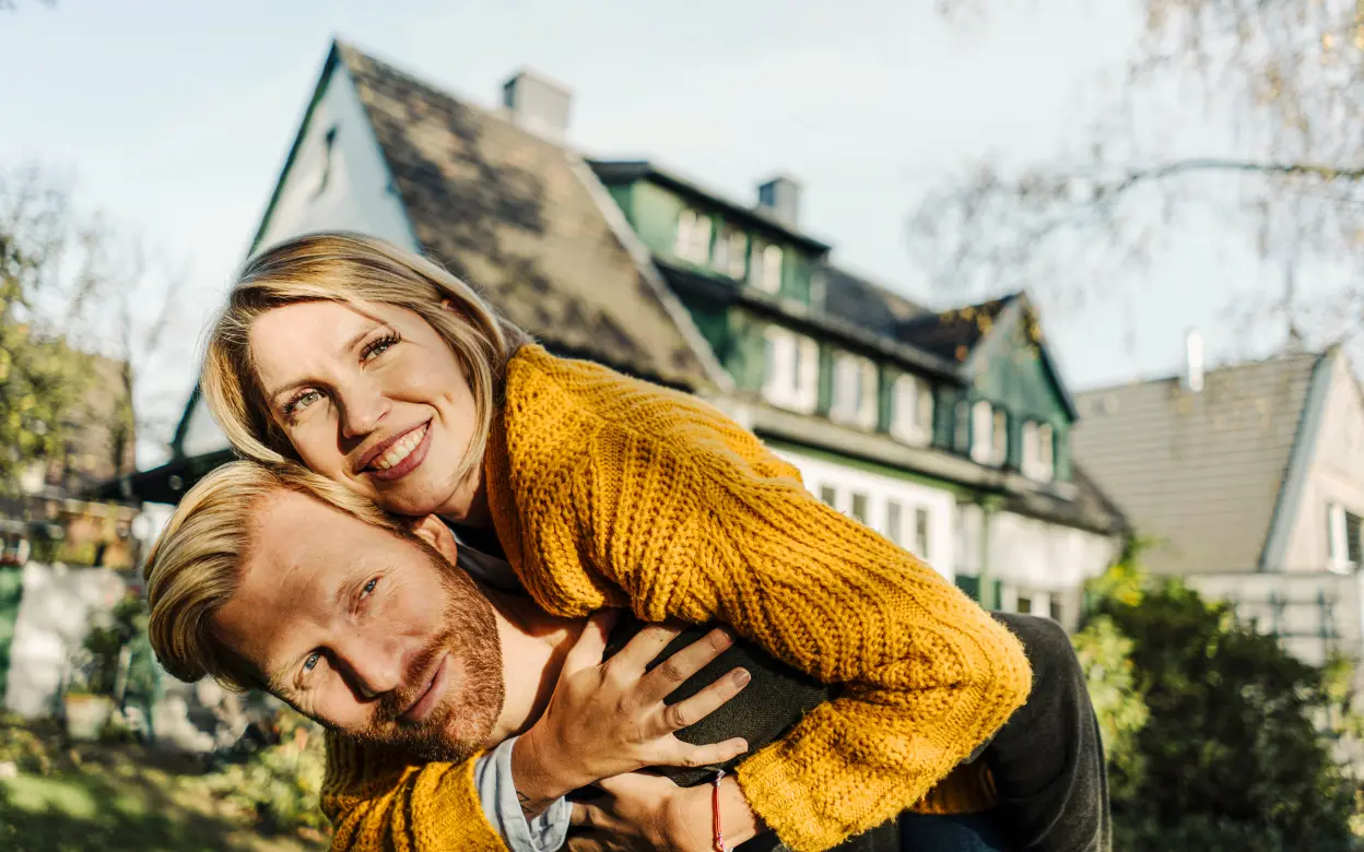Ein Mann trägt eine lächelnde Frau auf dem Rücken. Im Hintergrund steht ein Haus.