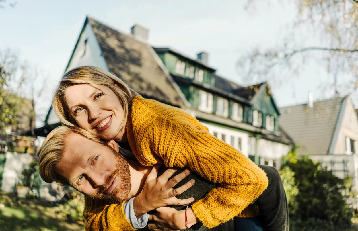 Ein Mann trägt eine lächelnde Frau auf dem Rücken. Im Hintergrund steht ein Haus.