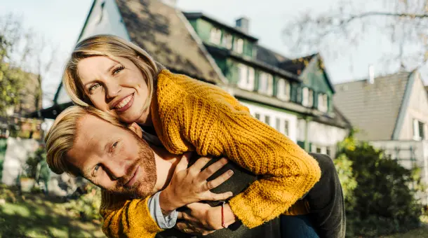 Ein Mann trägt eine lächelnde Frau auf dem Rücken. Im Hintergrund steht ein Haus.