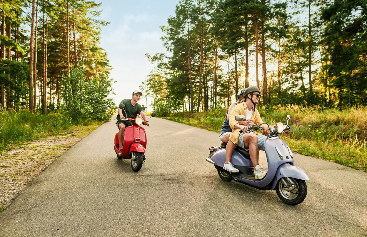 Zwei Rollerfahrer fahren fröhlich über eine Straße im Wald.