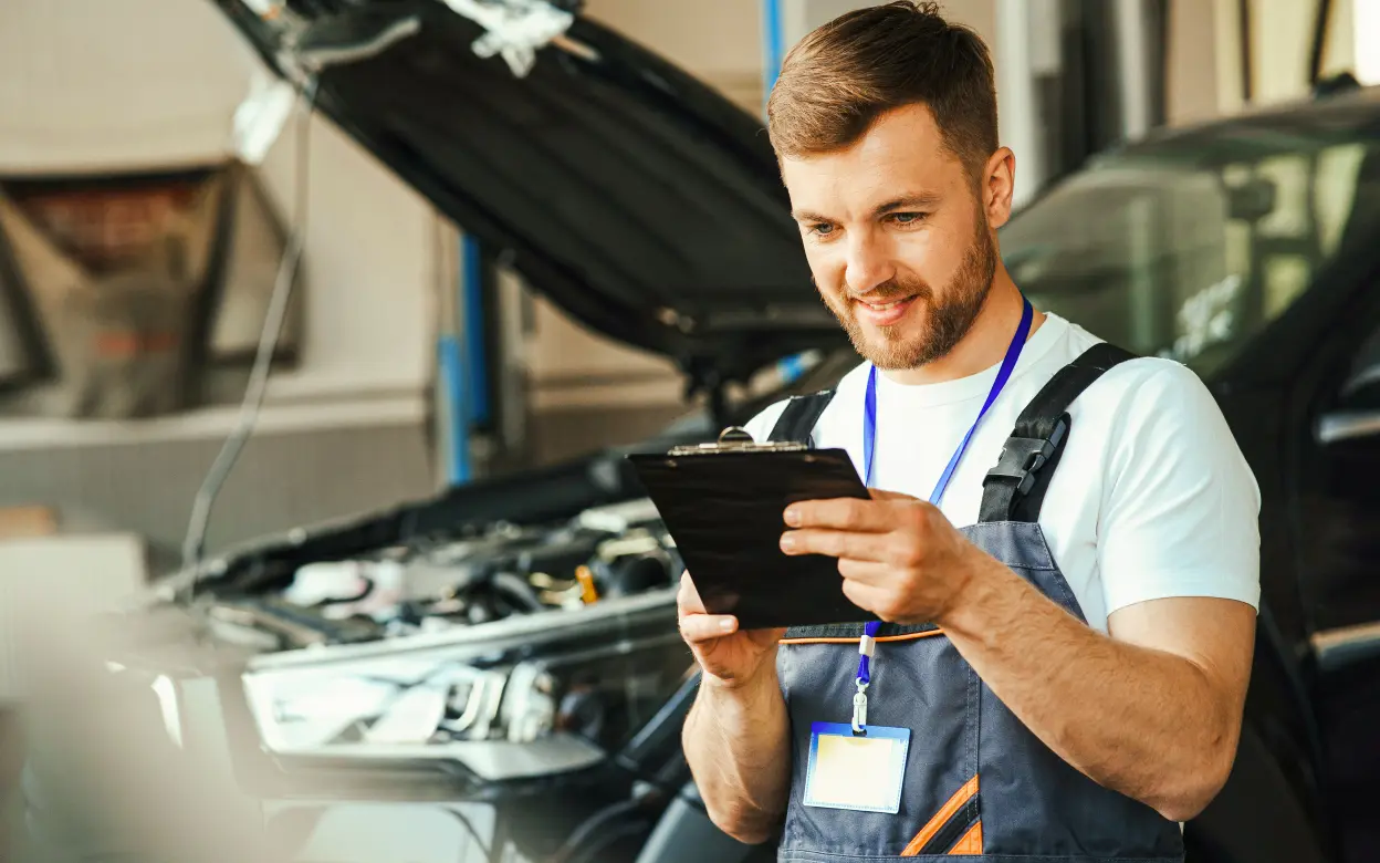 Ein Automechaniker steht vor einem Fahrzeug mit geöffneter Motorhaube und notiert etwas.