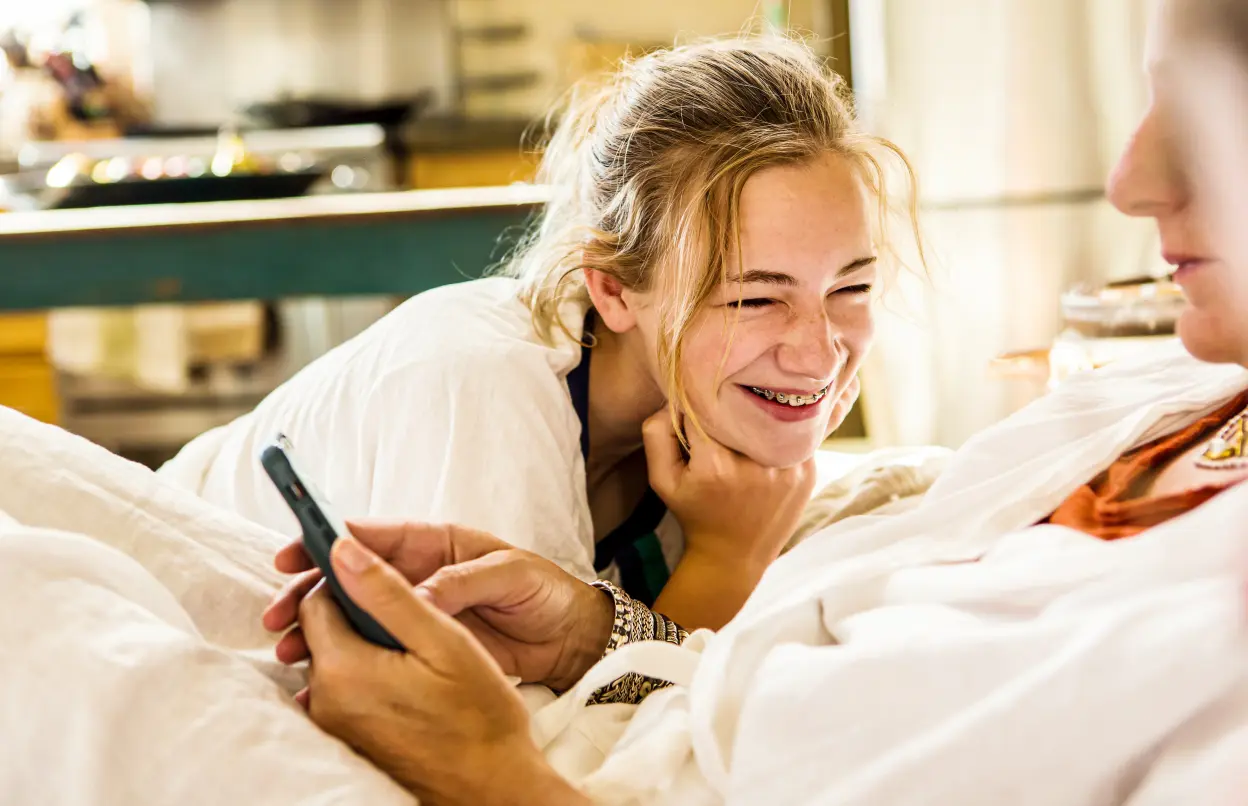 Ein Mädchen stützt sich lachend auf einem Bett neben einer liegenden Frau ab.