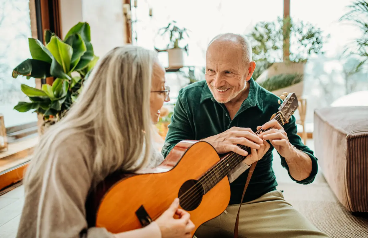 Ältere Dame wird von Ehemann beim Gitarre spielen unterstützt