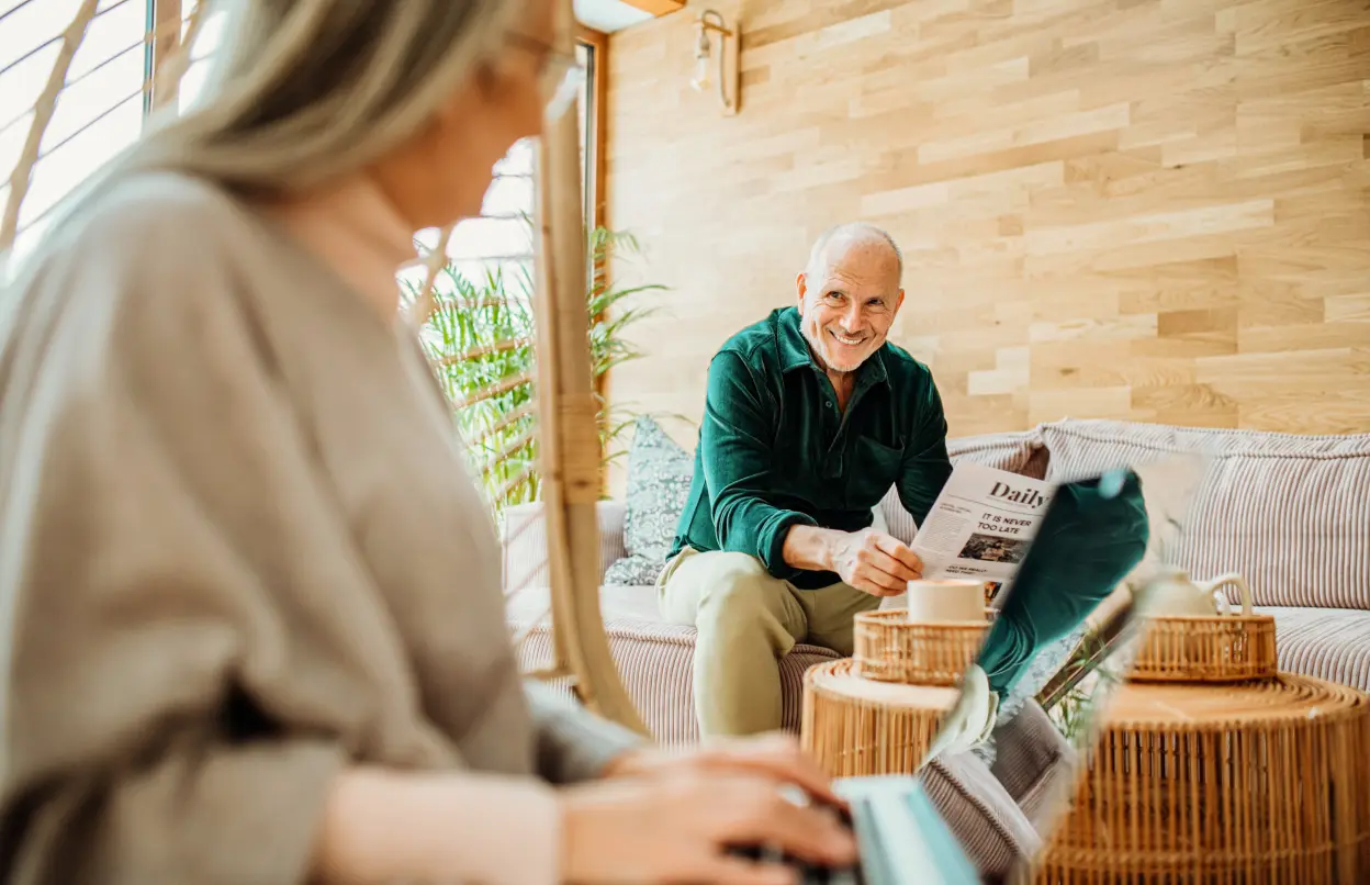 Ältere Frau sitzt am Laptop während Mann ihr zeitungslesend dabei zuschaut