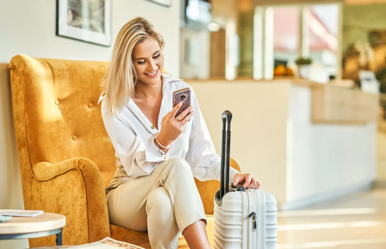 Frau sitzt mit Smartphone und Koffer in Hotellobby