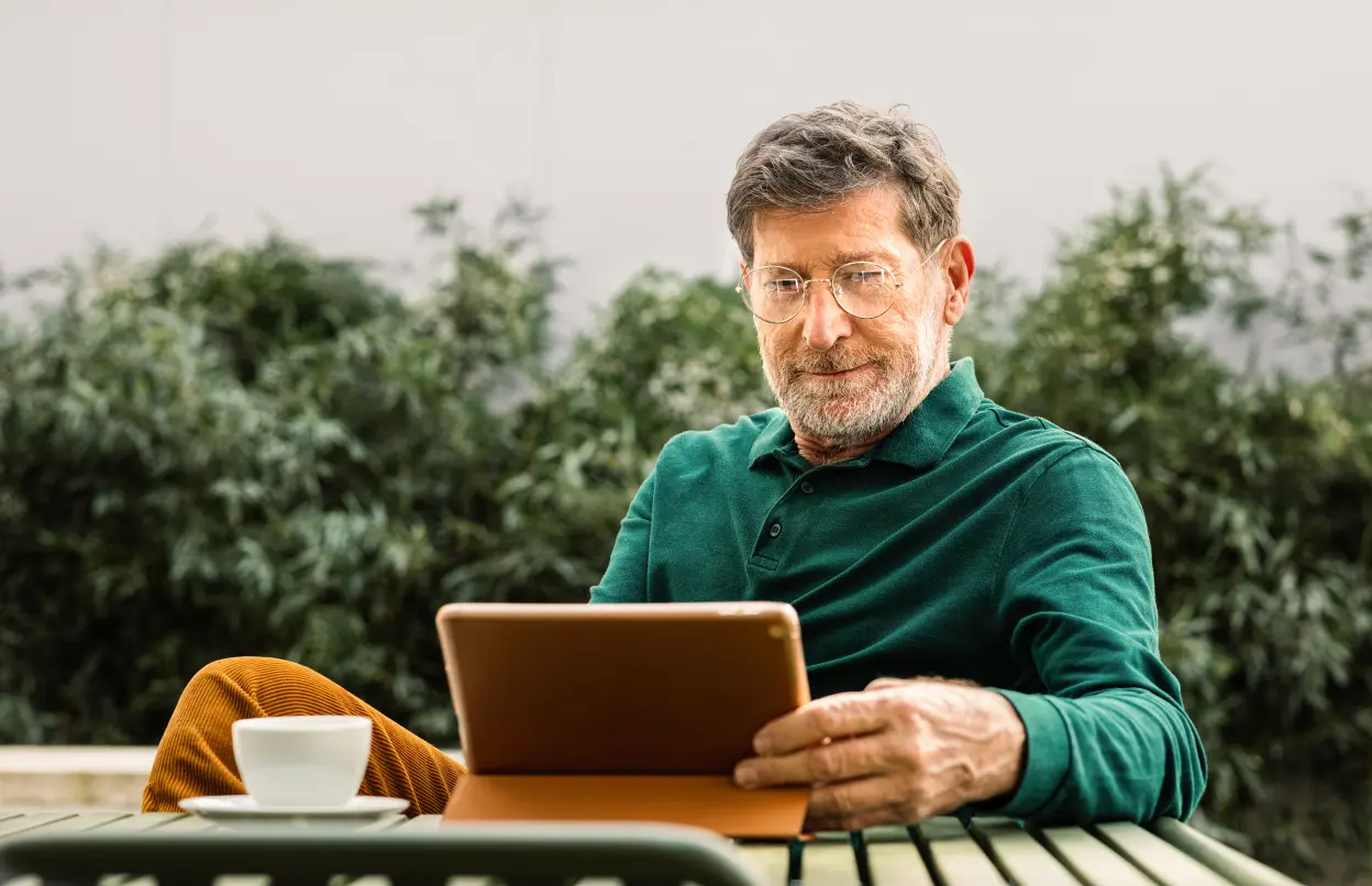 Ein Mann sitzt im Garten an einem Tisch mit einer Tasse und einem Tablet.