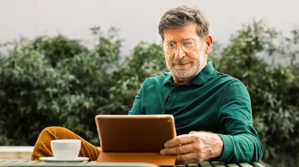Ein Mann sitzt im Garten an einem Tisch mit einer Tasse und einem Tablet.