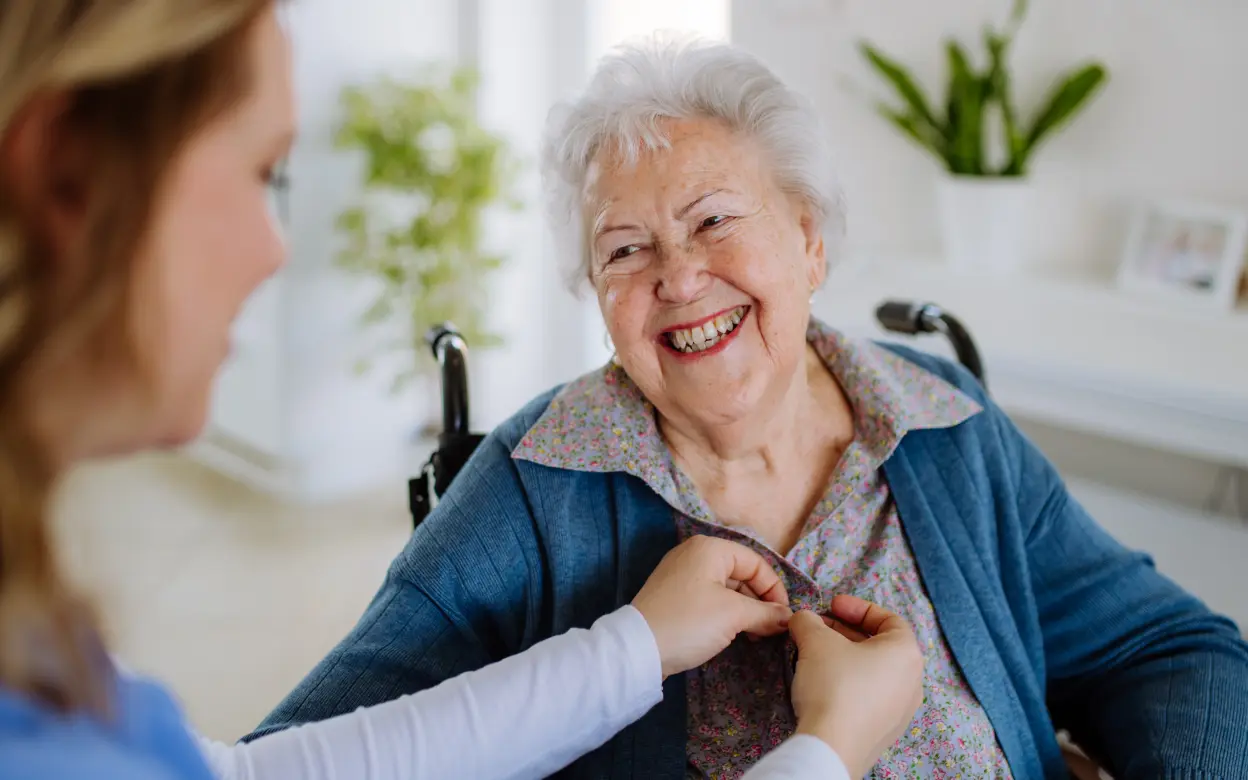Pflegerin hilft älterner Dame beim Zuknöpfen einer Bluse