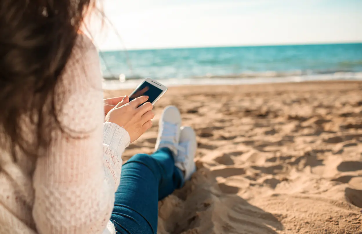 Eine Frau sitzt am Strand und tippt an ihrem Handy.