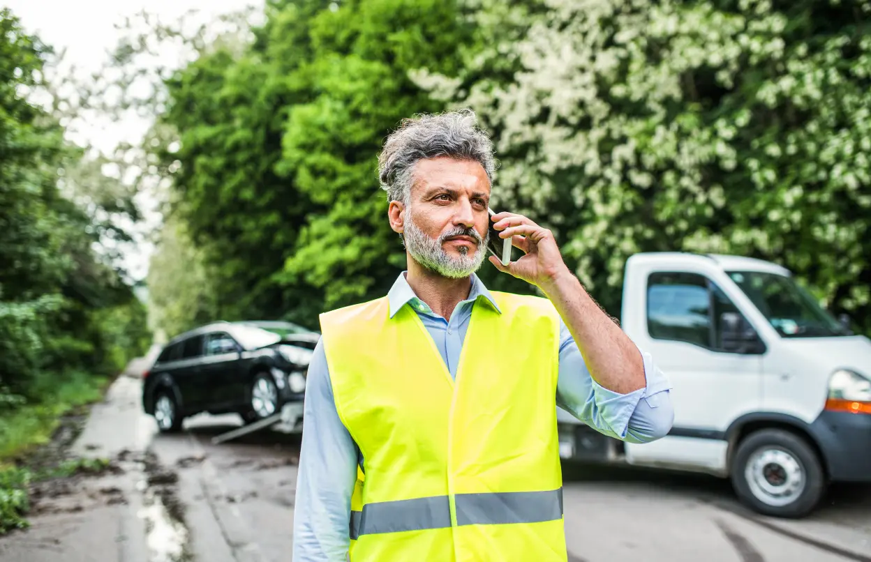 Eín Mann trägt eine Warnweste und telefoniert. Hinter ihm steht ein Abschleppwagen.