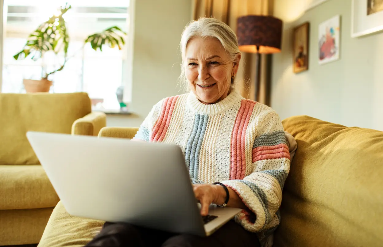 Eine Frau sitzt auf einem Sofa und arbeitet lächelnd am Laptop.