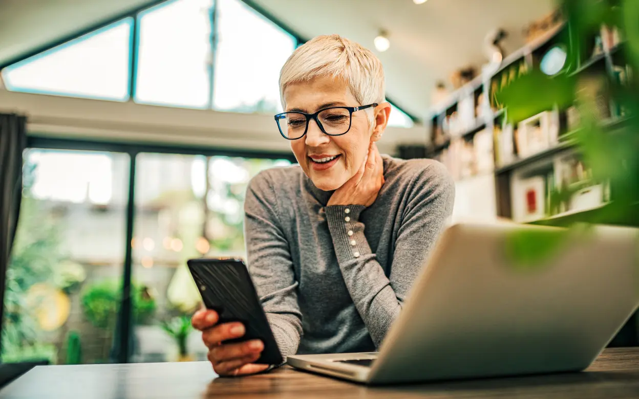 Eine Frau sitzt an einem Laptop und schaut lächelnd auf ihr Handy.